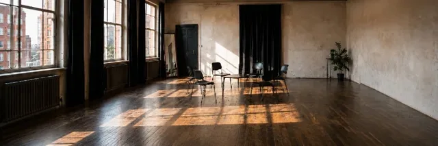 Sunlit acting studio with a circle of black chairs around a low table, wooden floors and large windows